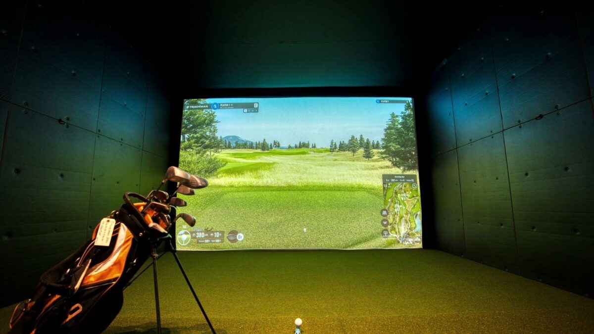 Indoor golf simulator bay with a course on screen and a golf bag on the turf