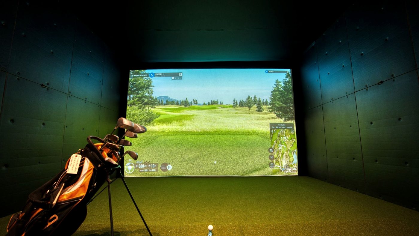 Indoor golf simulator bay with a course on screen and a golf bag on the turf