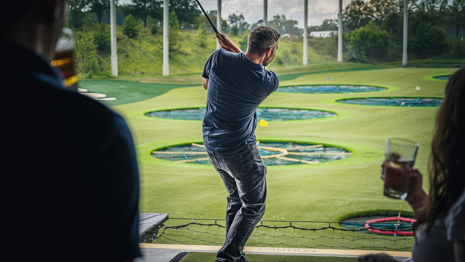 Friends with drinks watching a man swing at an entertainment driving range bay