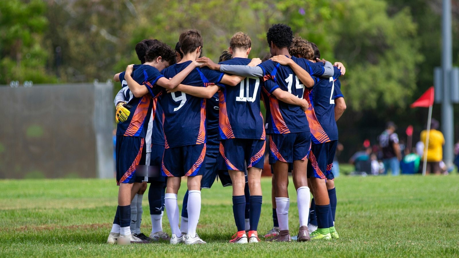Youth soccer team in a tight huddle, arms around each other, on a grass field