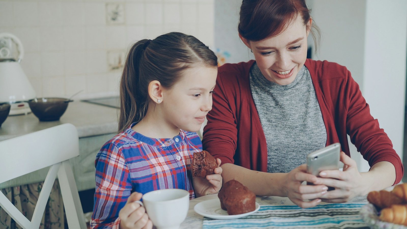 Mother scrolling her phone at the kitchen table beside her daughter